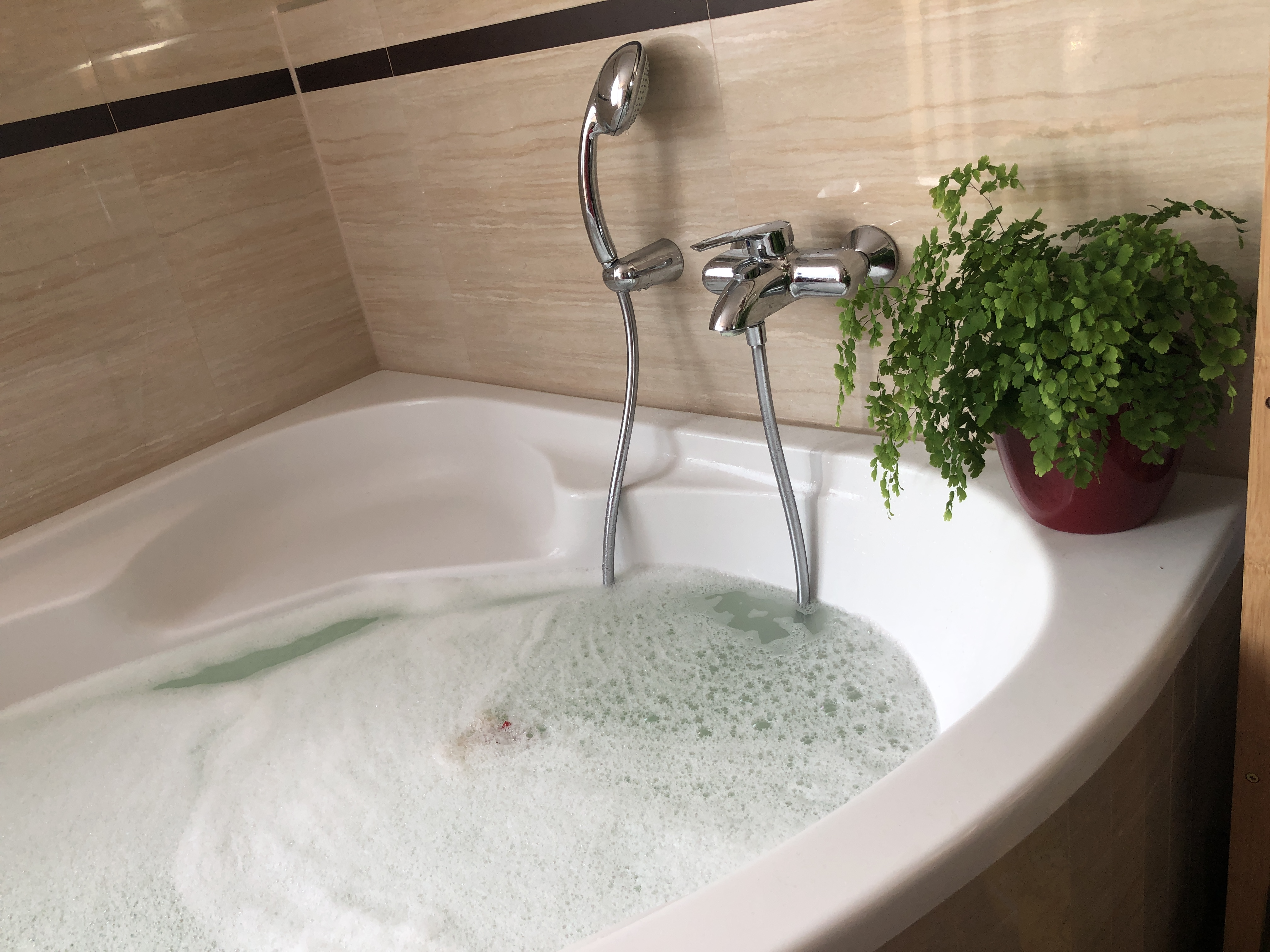 A corner bath with a light layer of bubbles, an oatmeal scrubby bag, and a potted fern on one corner say 'relax'.
