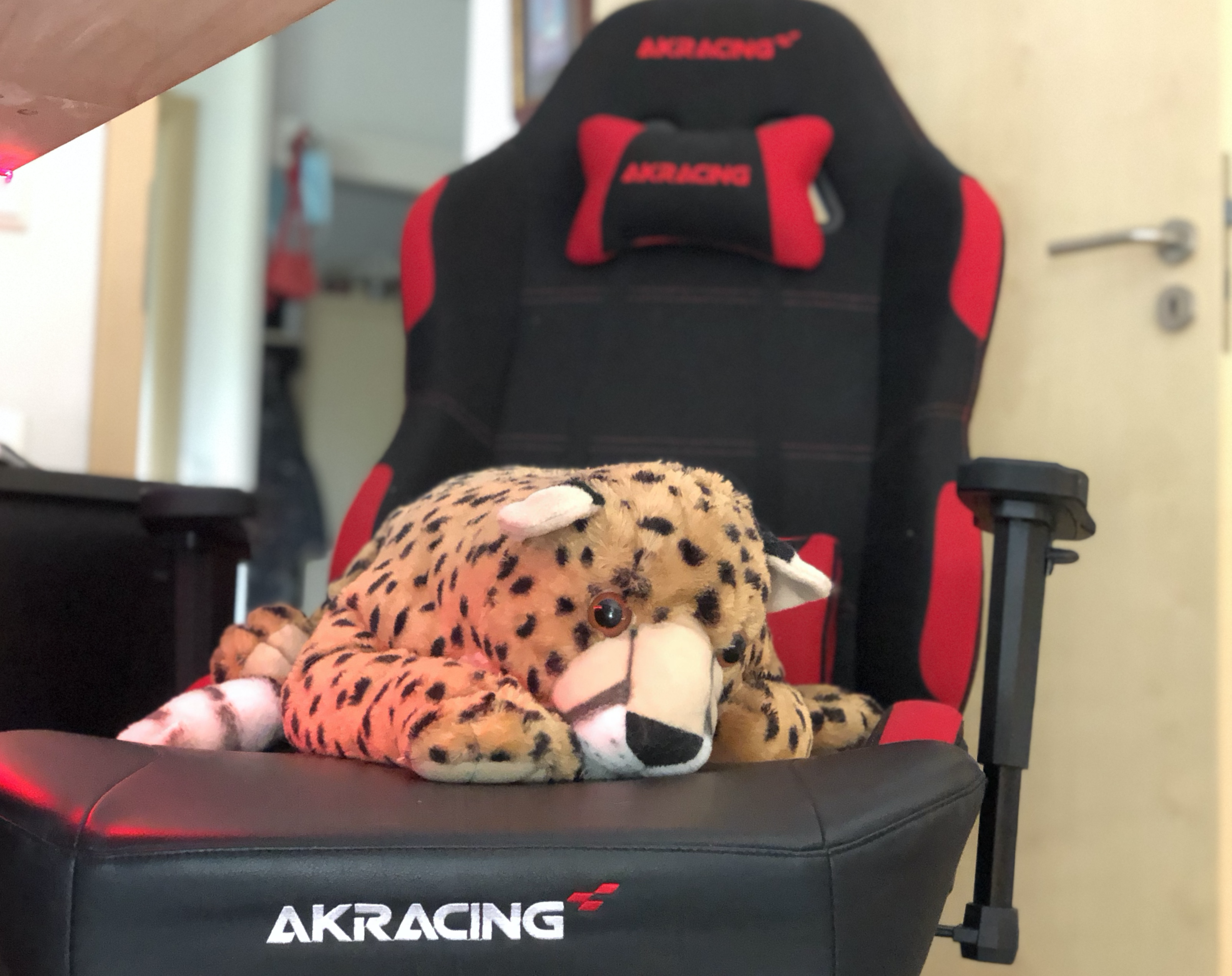 A large soft toy of an Amur leopard lies tiredly on a red and black AKRacing gaming chair in a home office