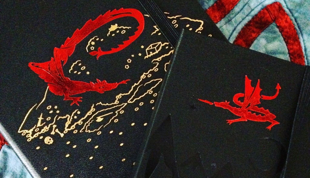 Two black Moleskin journals from the Lord of the Rings series, both with red dragons on the front - one dragon curled on top of a pile of gold and the other flying over black embossed mountains
