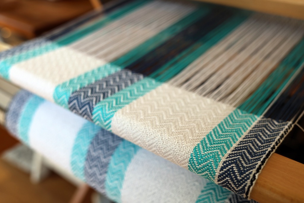 A handwoven twill placemat in progress on a table loom in white, denim blue and turquoise.