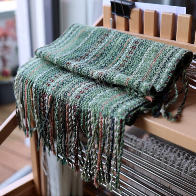A green handwoven woollen scarf is folded on the table loom with its twisted fringe hanging down