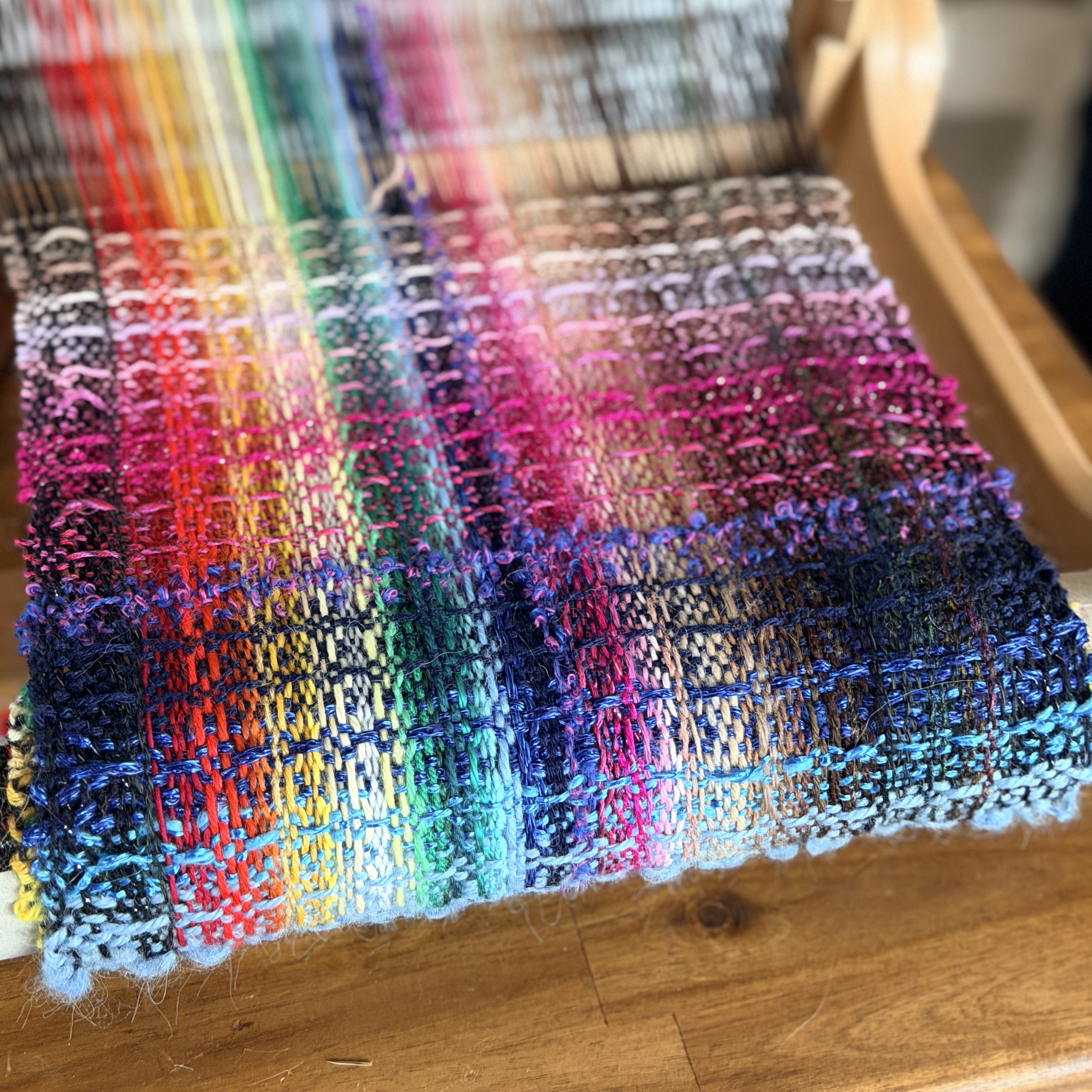 A rainbow scarf of all different types of yarn is being woven in a waffle pattern on a rigid heddle loom